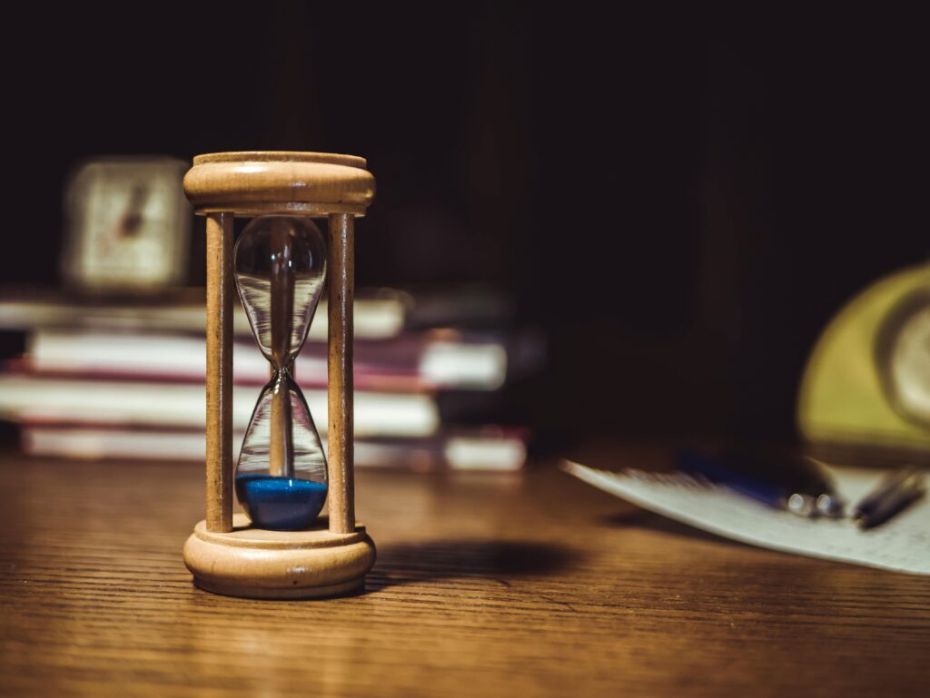 A close-up of a wooden hourglass with blue sand on a wooden desk, symbolizing time and patience.